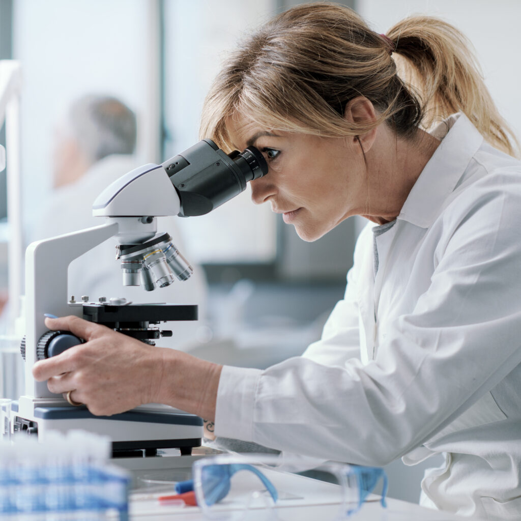 Vrouw met witte jas kijkt in microscoop